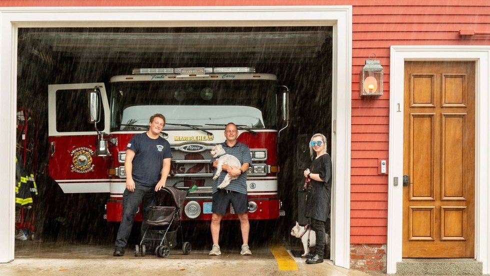 Firefighters and a neighbor with their dogs shelter from rain in an open firehouse engine bay.